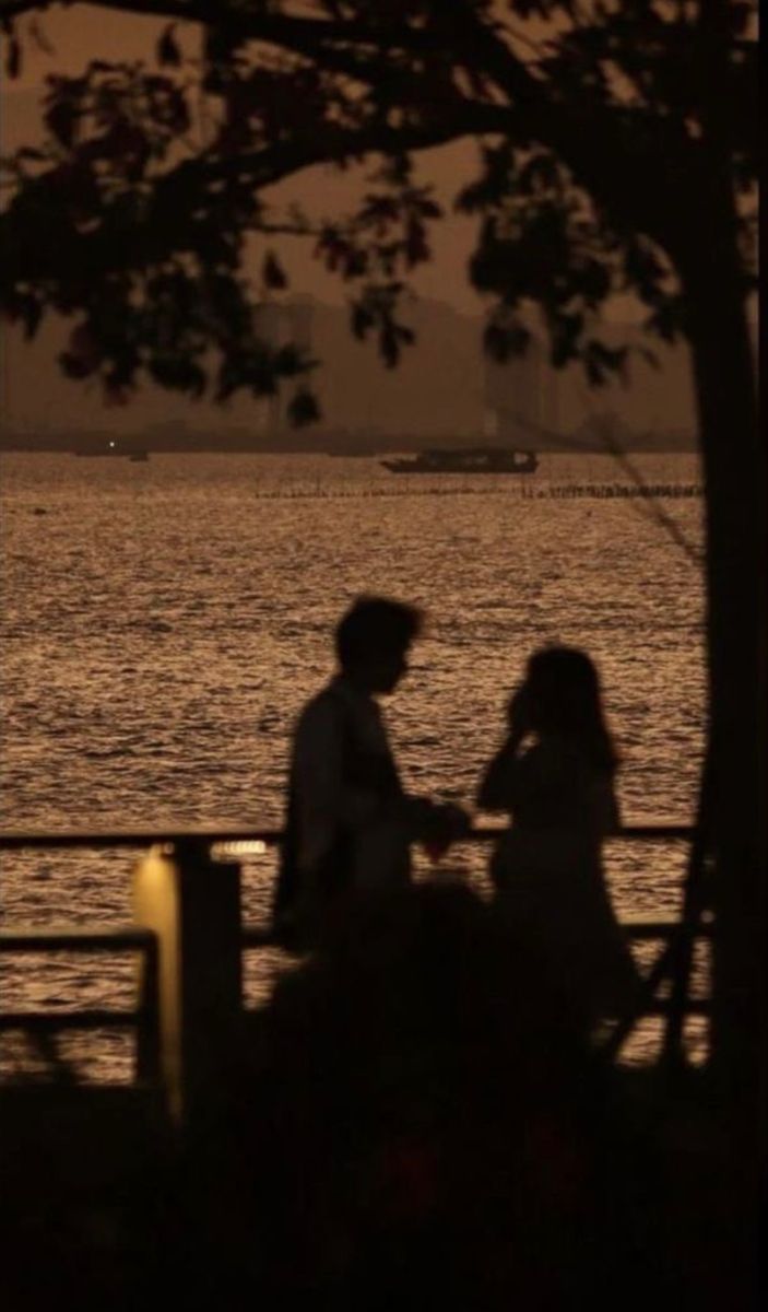 two people sitting on a bench next to the water at sunset with boats in the distance