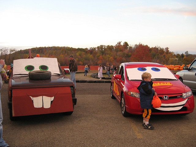 a young boy standing next to a red car in front of other cars and people