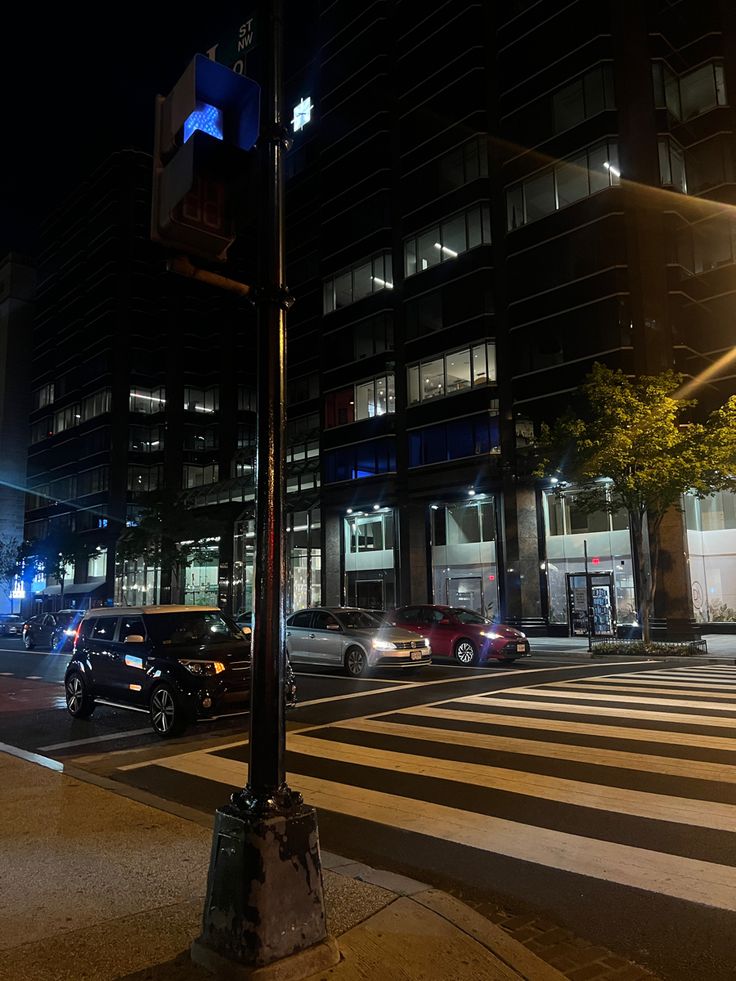 an intersection at night with cars driving on the road and tall buildings in the background