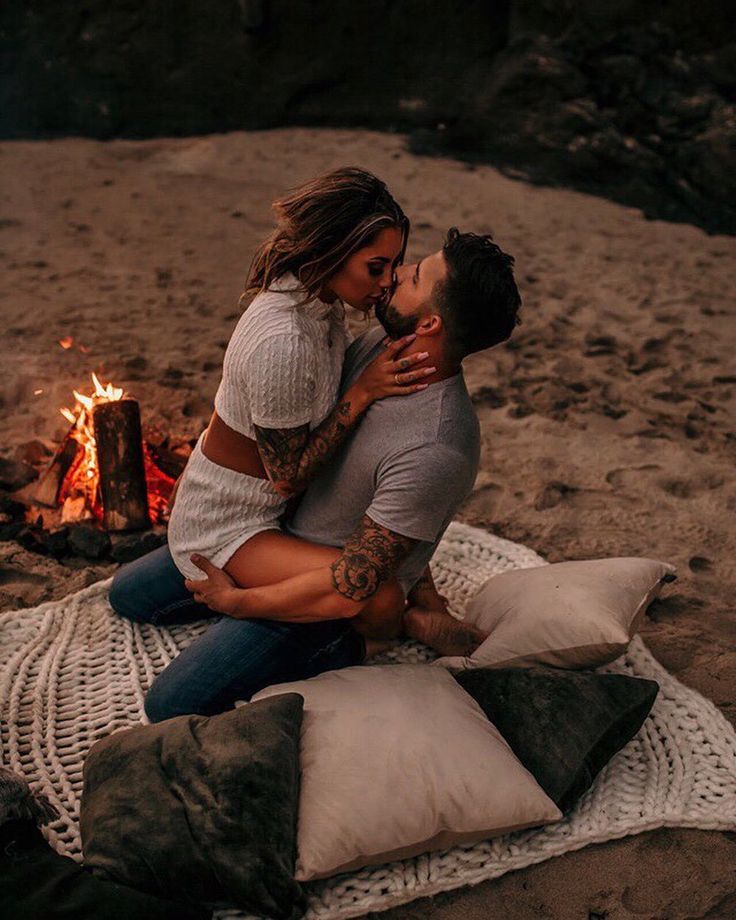 a man and woman sitting next to each other on a blanket in front of a campfire