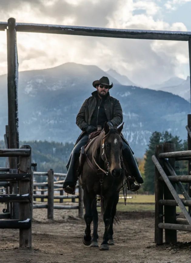 a man riding on the back of a brown horse next to a wooden fence and mountains