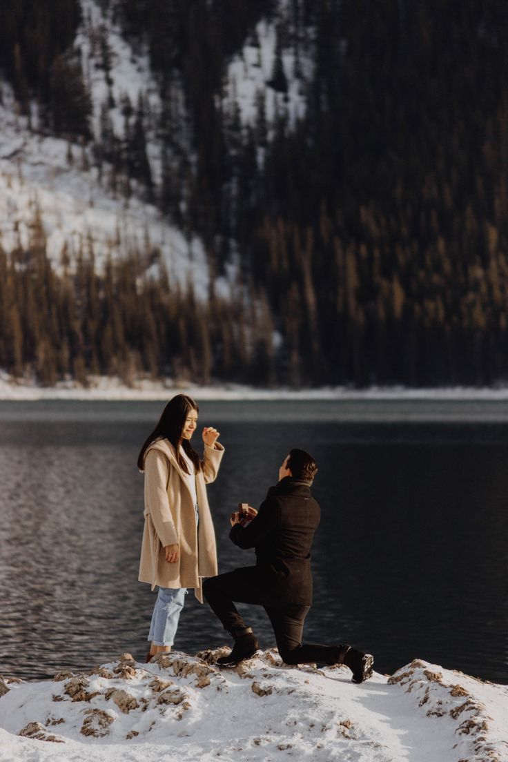 a man kneeling down next to a woman on top of a snow covered ground near a lake