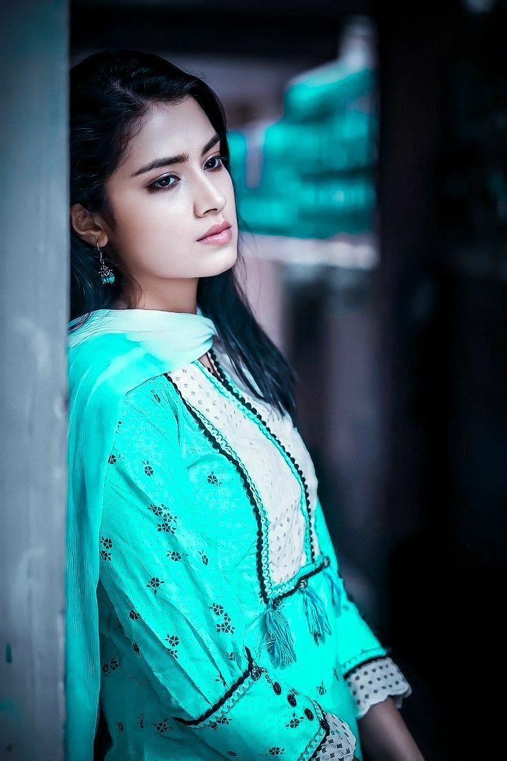 a beautiful young woman standing next to a wall wearing a blue dress and jewelry on her neck