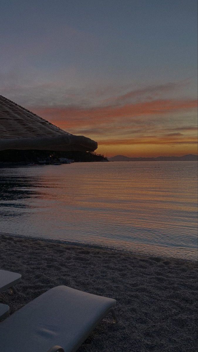 two lounge chairs sitting on top of a sandy beach next to the ocean at sunset