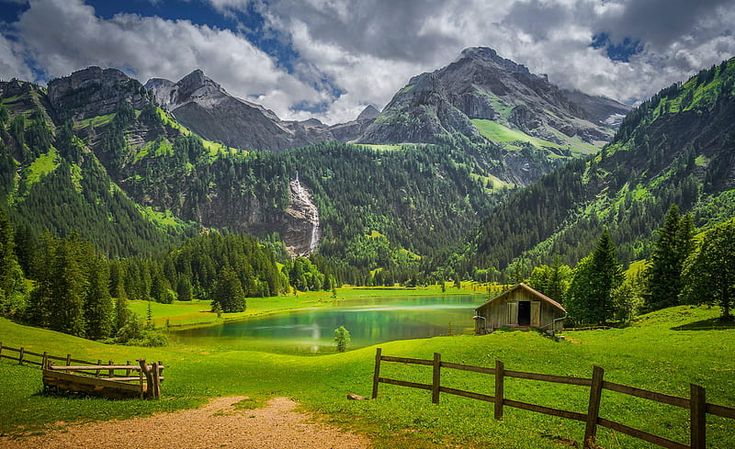 there is a small cabin in the middle of this mountain meadow with a lake and mountains behind it