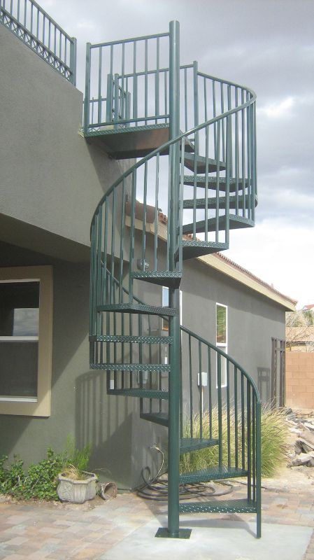 a metal spiral staircase in front of a gray building with green railings on the sides