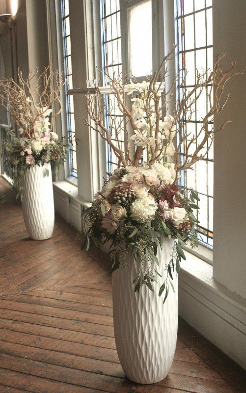 two white vases with flowers and branches in them on a wooden floor next to windows