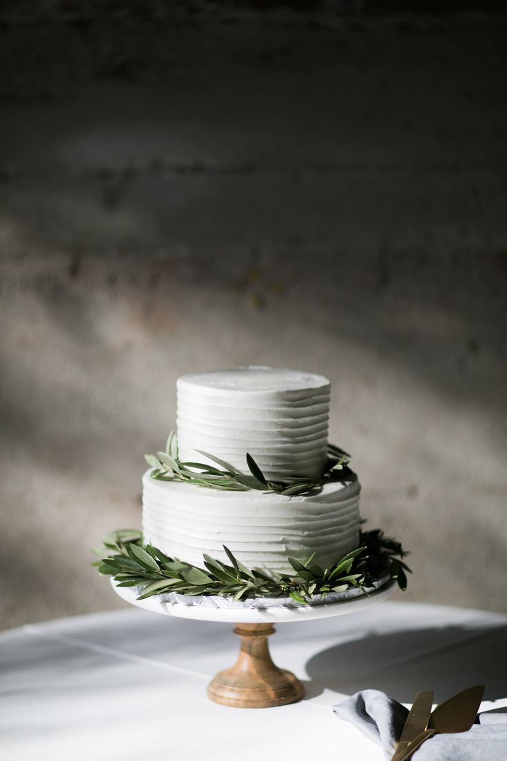 a three tiered cake with greenery on top sits on a white tablecloth