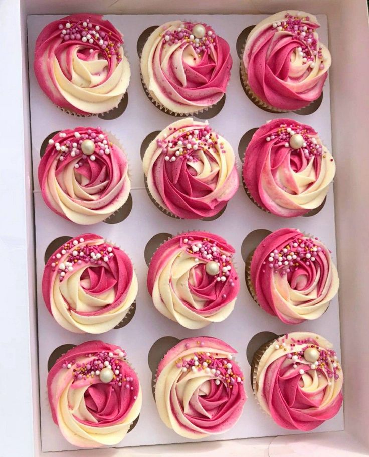 twelve cupcakes with pink and white frosting in a box on a table