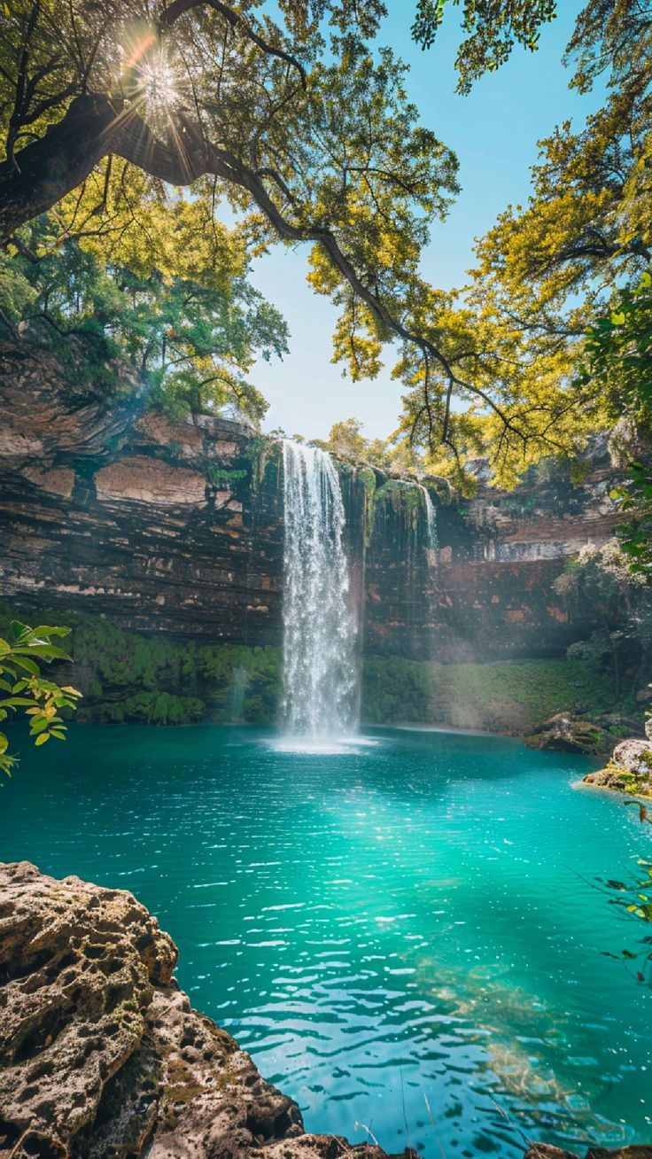 a waterfall in the middle of a lake surrounded by trees and rocks with blue water