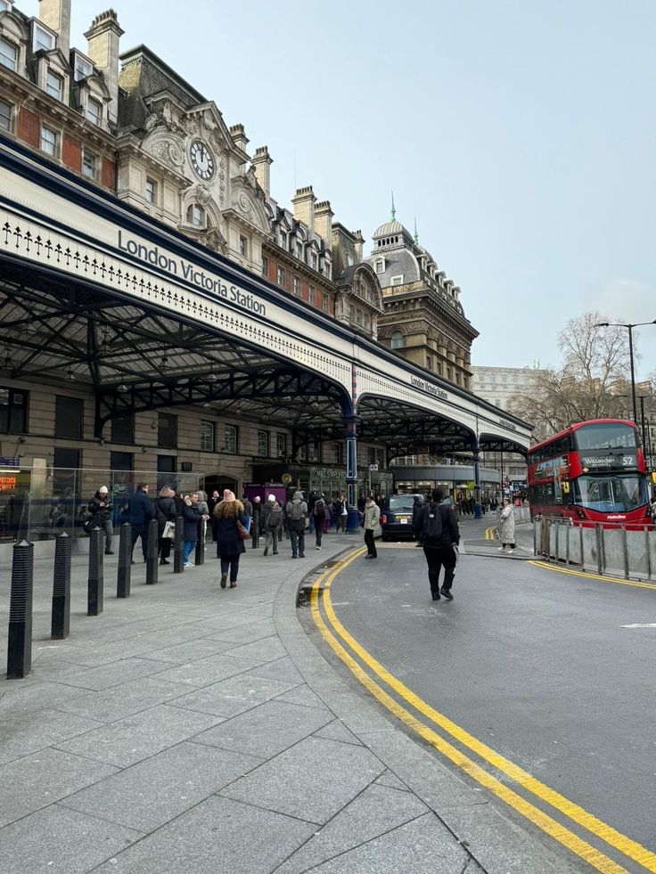 people are walking on the sidewalk in front of a train station with two red double decker buses