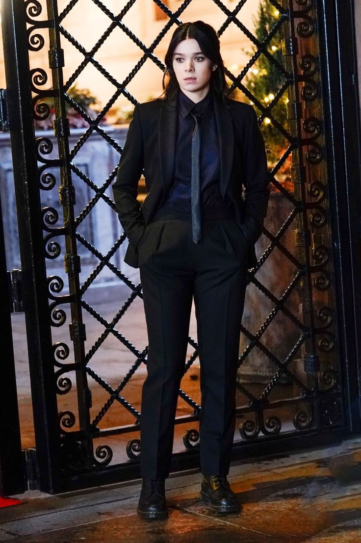 a woman standing in front of an iron gate wearing a suit and tie with her hands on her hips