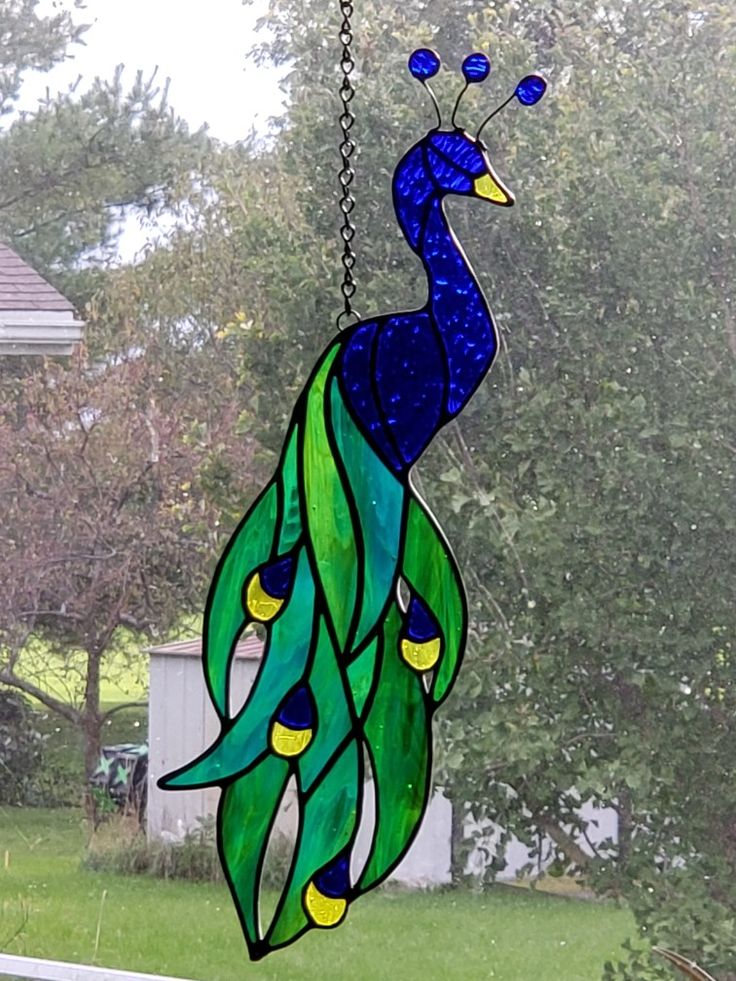 a stained glass peacock hanging from a chain on a window sill in front of a house