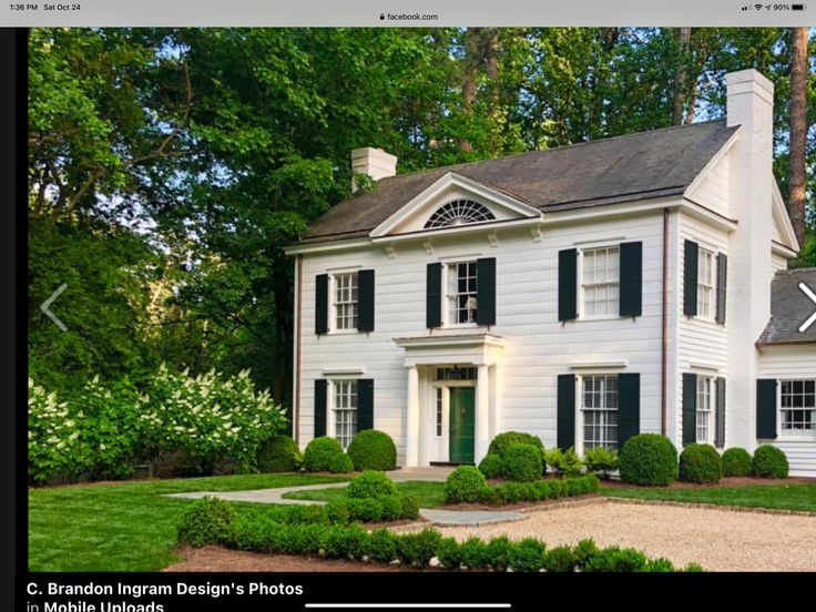 a large white house with black shutters on the front and side windows, surrounded by lush green trees