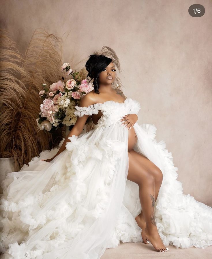 a woman in a white dress sitting on a chair with her legs crossed and flowers behind her