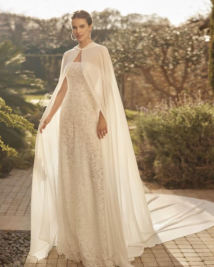 a woman in a white gown and cape standing on a brick walkway with trees behind her