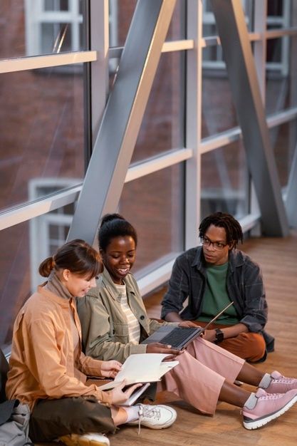 three young people sitting on the floor in front of a window looking at their laptops