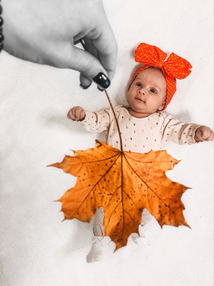 a baby laying on the ground with a leaf in front of her face and a hand reaching for it