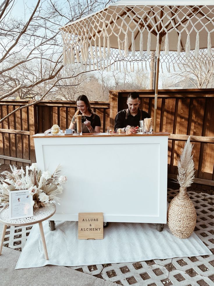 two people sitting at an outdoor bar on a patio with white rugs and wooden tables