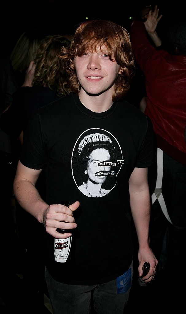 a young man standing in front of a crowd holding a drink and wearing a black t - shirt