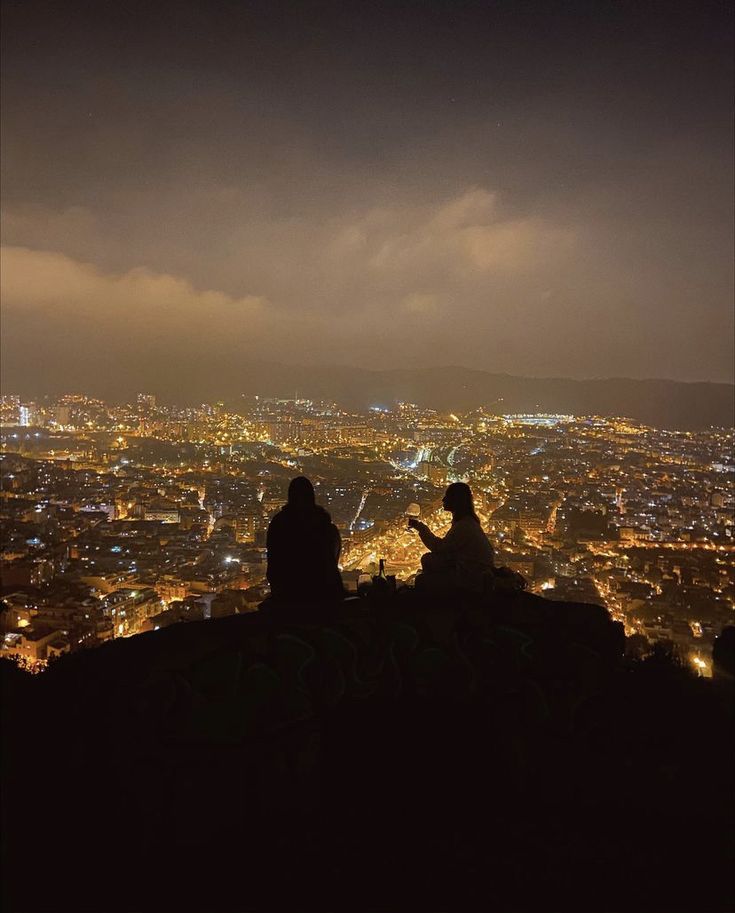 two people sitting on top of a hill looking at the city lights in the distance