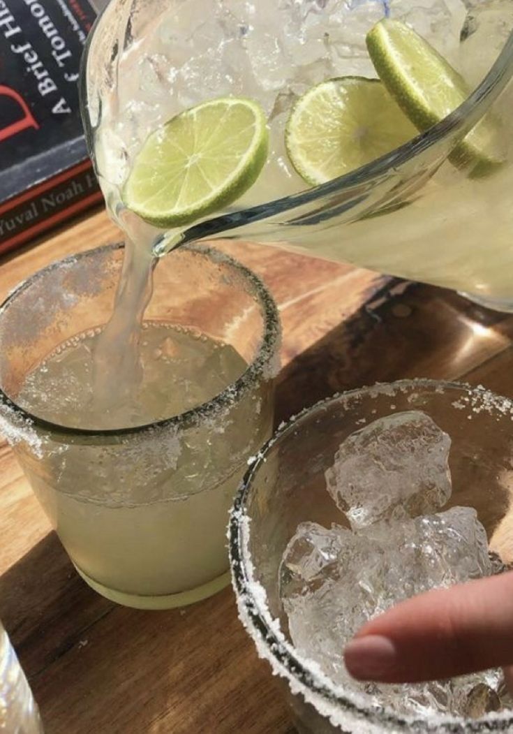 two glasses filled with ice and limes next to a book on a wooden table