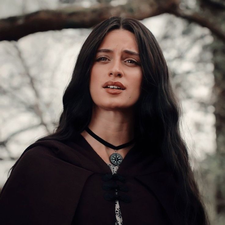 a woman with long black hair wearing a brown cape and necklace standing in front of a tree