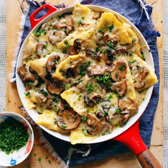 a red skillet filled with pasta covered in mushrooms and parsley on top of a blue cloth