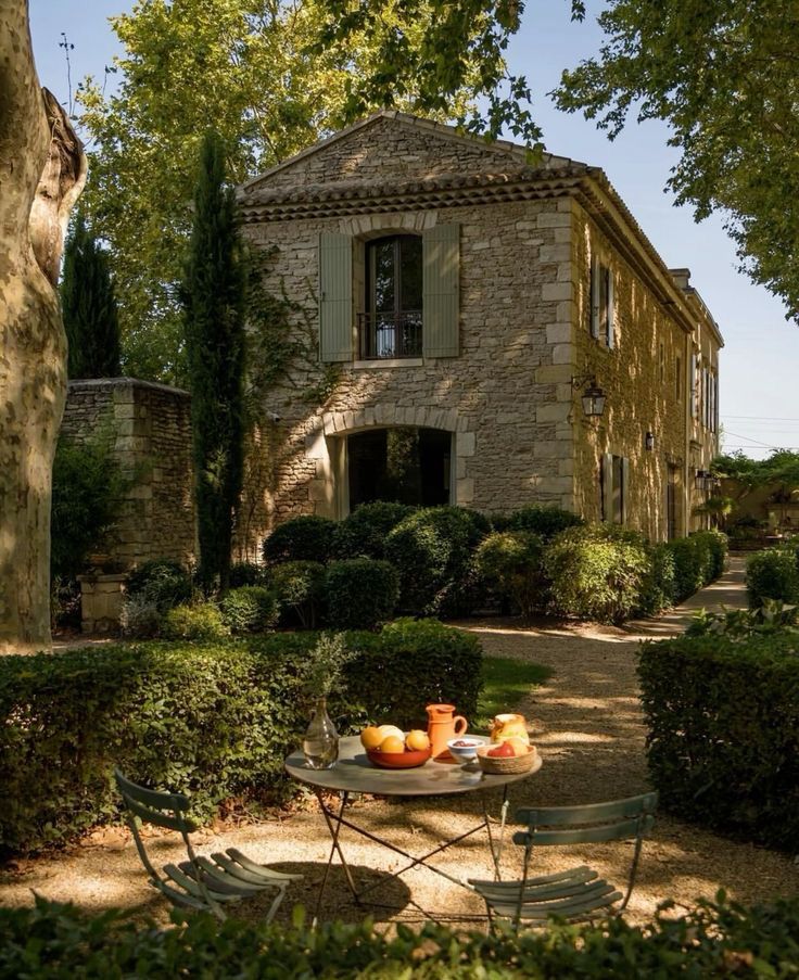 an outdoor table with fruit on it in front of a stone house surrounded by bushes and trees