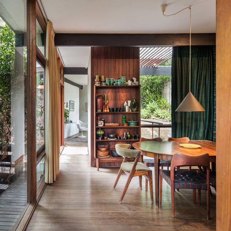 a dining room with wooden floors and green curtains on the window sill, next to an open bookcase filled with books