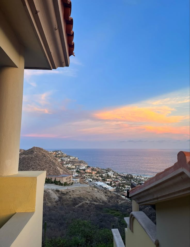 the view from an apartment balcony at sunset overlooking the ocean and town below, with mountains in the distance