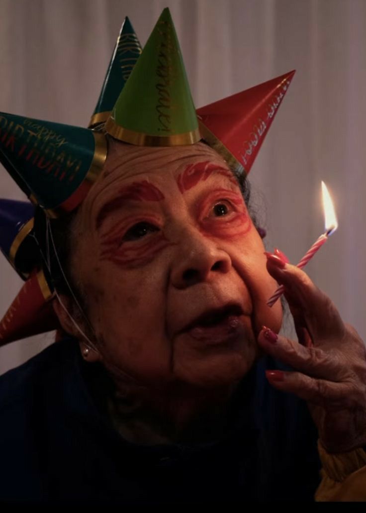 an old woman wearing party hats and holding a lit match between her fingers to her face