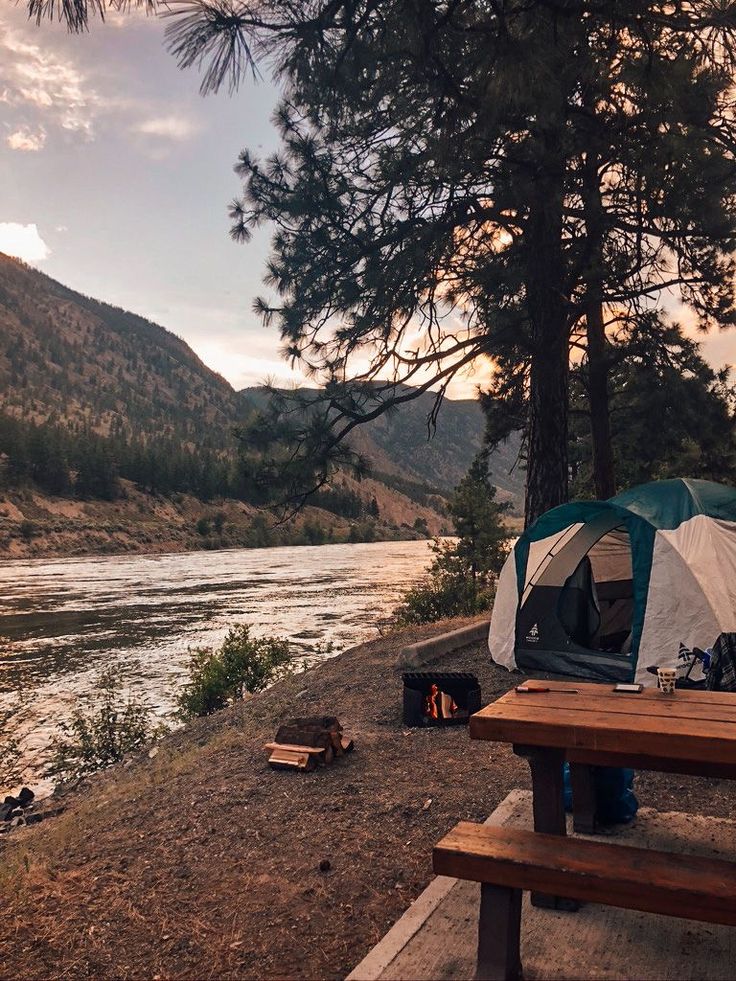 a tent set up next to a river with mountains in the background and picnic tables