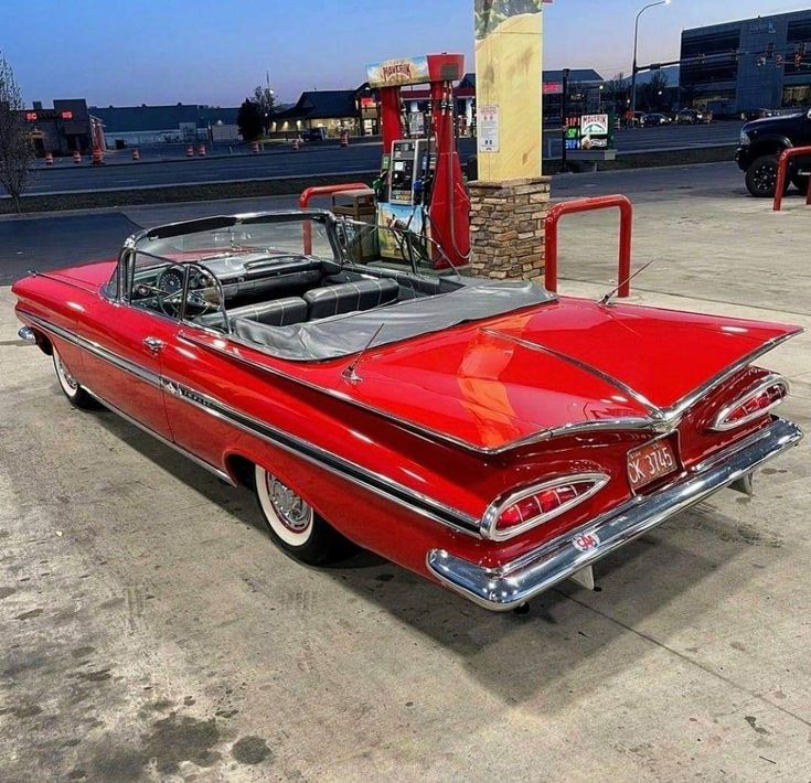 an old red car parked in front of a gas station with its hood up and the top down