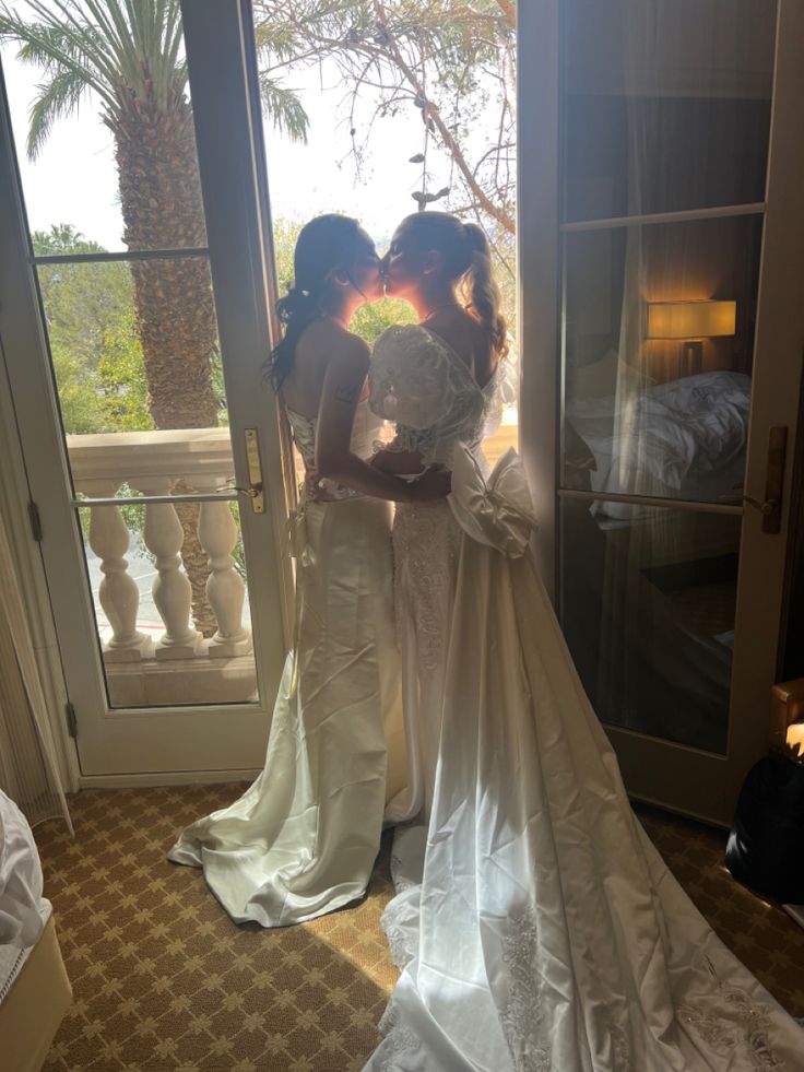 two brides standing in front of a window with their arms around each other and kissing