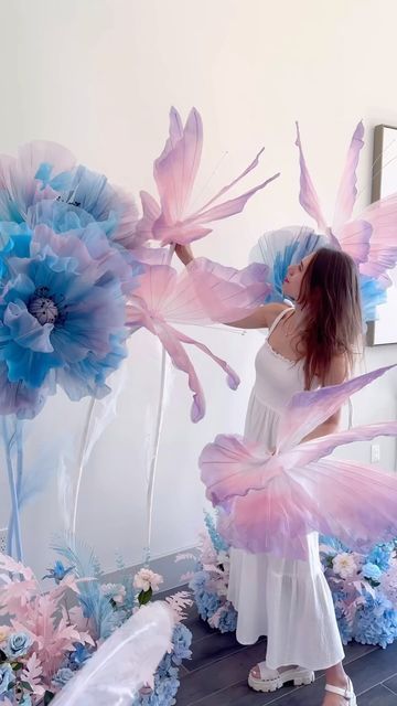 a woman in white dress standing next to large blue and pink flowers with wings on them