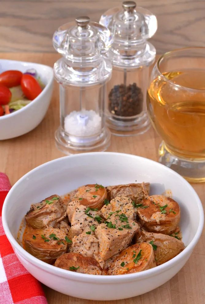 a white bowl filled with meat and vegetables next to two glasses of wine on a wooden table