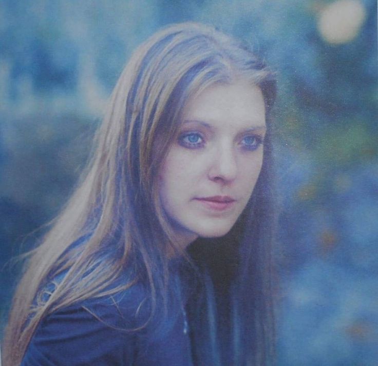 a woman with long hair and blue eyes is posing for a photo in front of trees