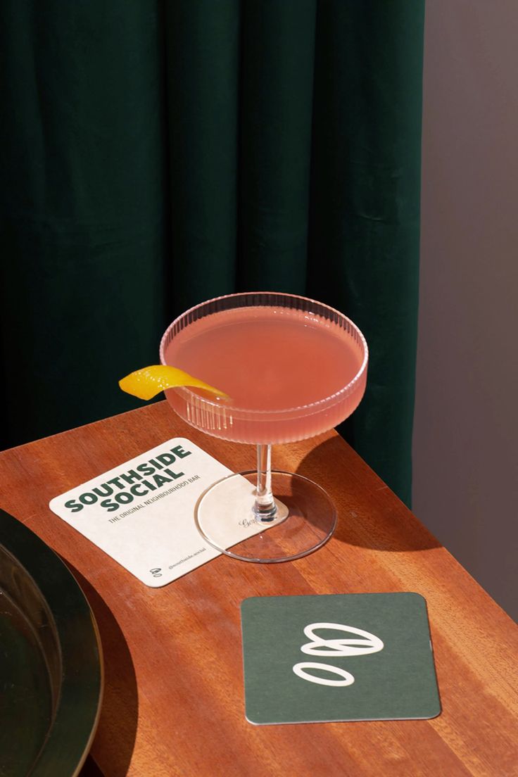 a pink drink sitting on top of a wooden table next to a black plate and green curtain