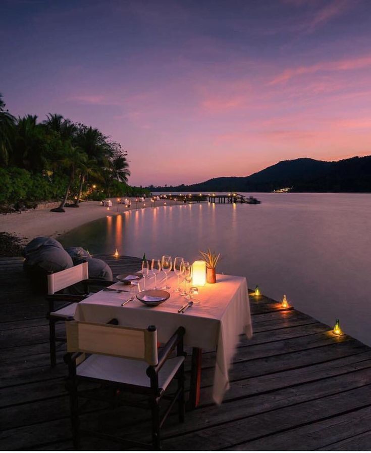 a dinner table set up on a dock with candles and wine glasses in front of it