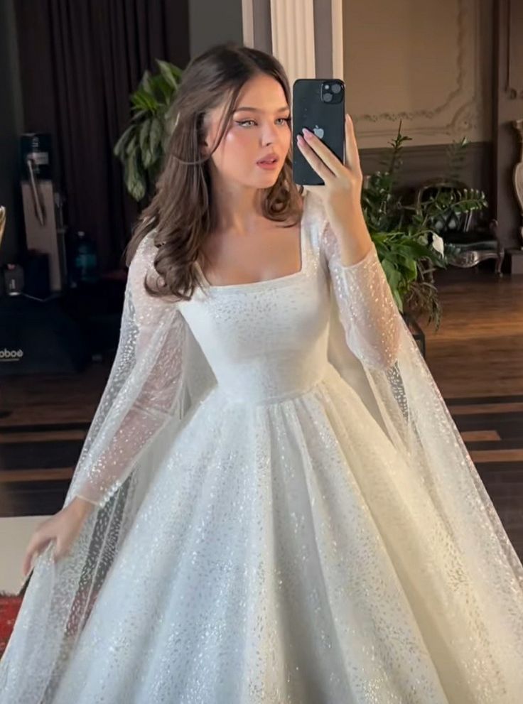 a woman in a white wedding dress taking a selfie with her cell phone while wearing a long sleeved gown