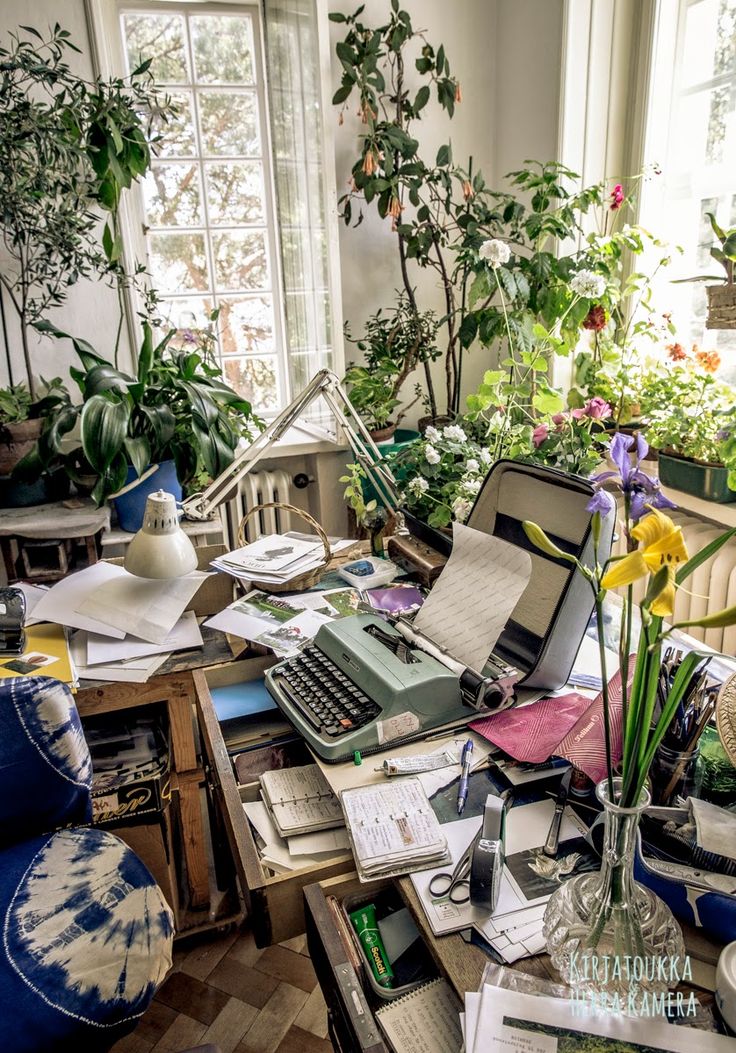 a cluttered desk with an old typewriter on it and lots of plants in the background