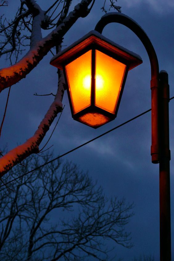 a street light is lit up in the night sky with snow on the ground and bare trees behind it