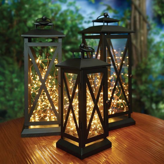 two lanterns with fairy lights on them sitting on a table in front of a forest