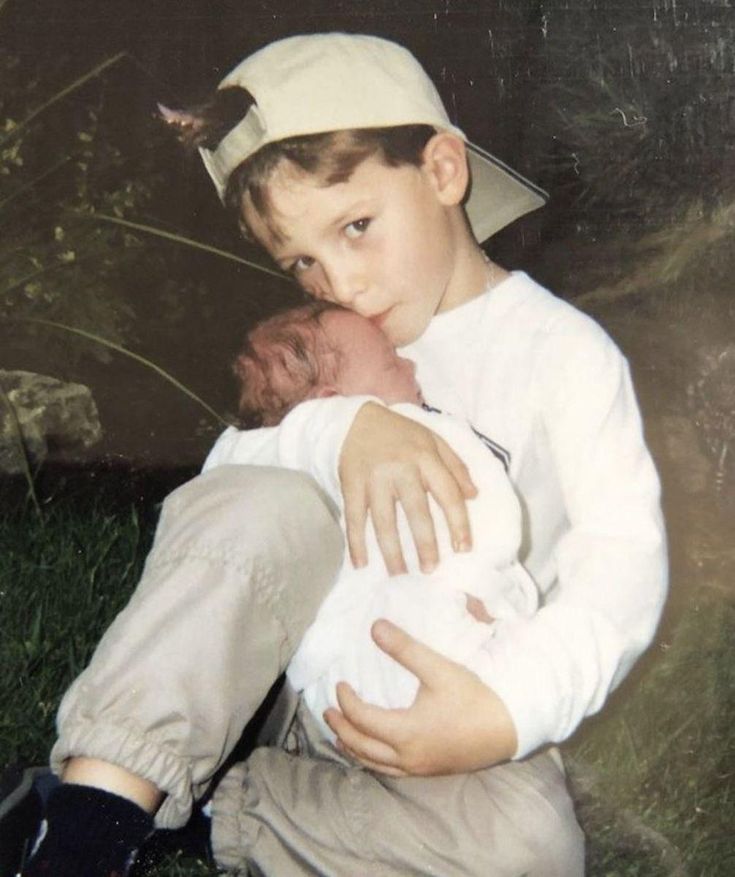 a young boy sitting on the ground holding a baby while wearing a baseball cap and white shirt