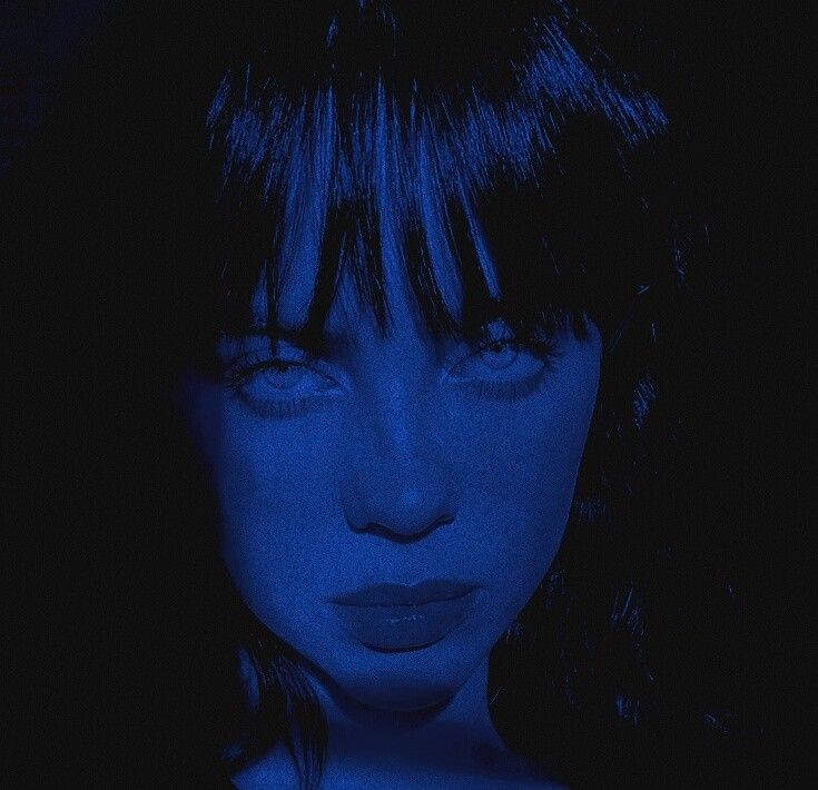 a woman with dark hair and blue makeup looks at the camera while she's illuminated