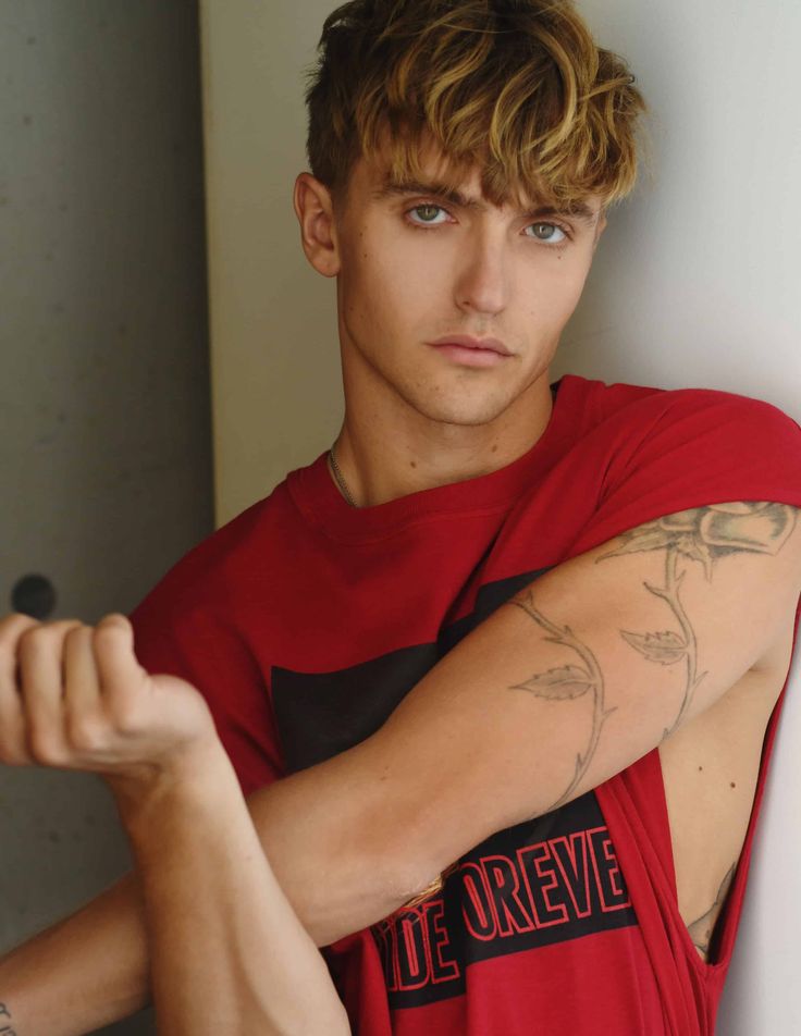 a young man in a red shirt is leaning against a wall with his arms crossed