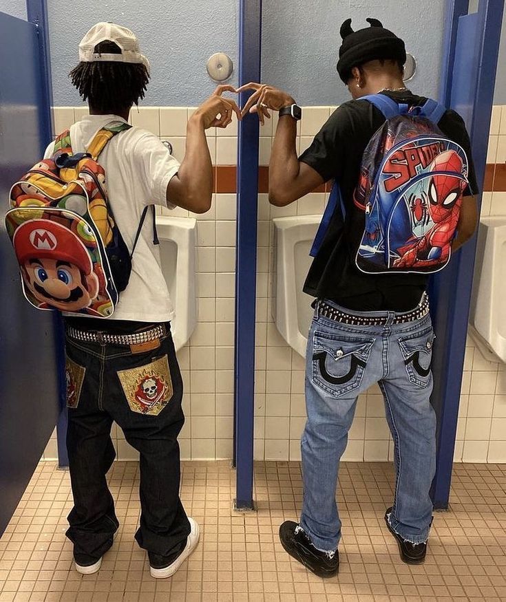 two boys with backpacks standing in front of urinals and one boy is holding an object