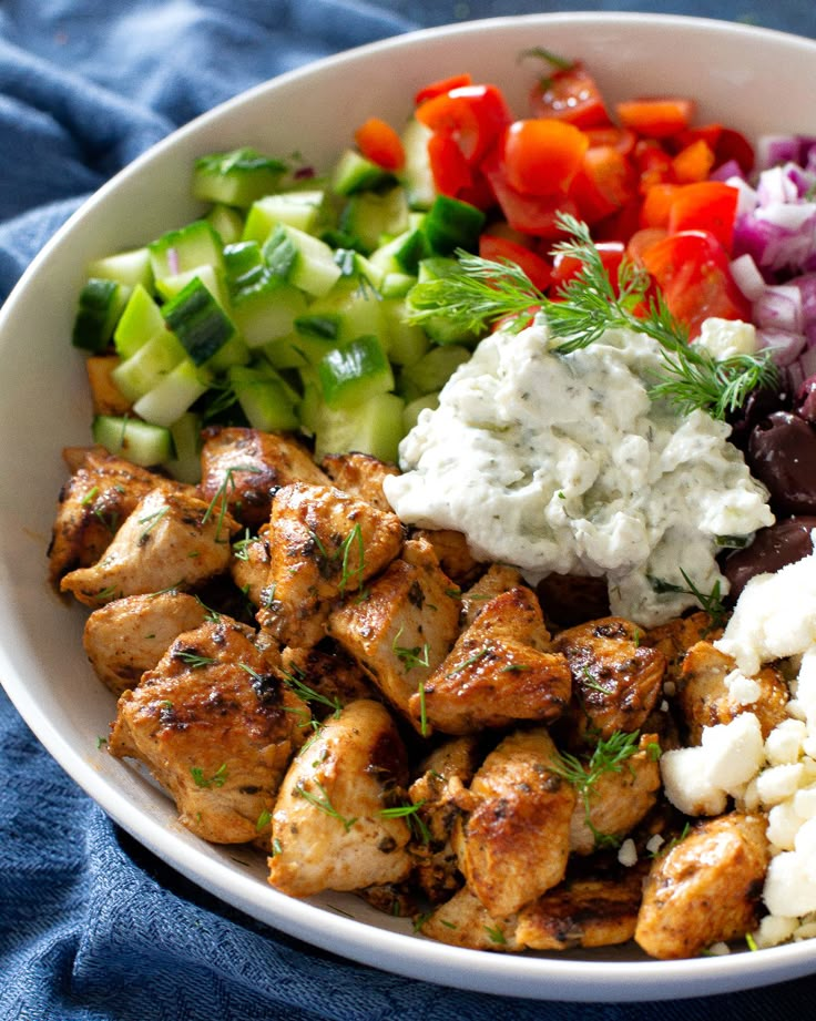 a white bowl filled with chicken, vegetables and feta cheese on a blue cloth