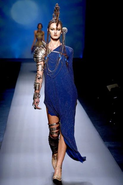 a woman walking down a runway in a blue dress with chains on her arms and legs